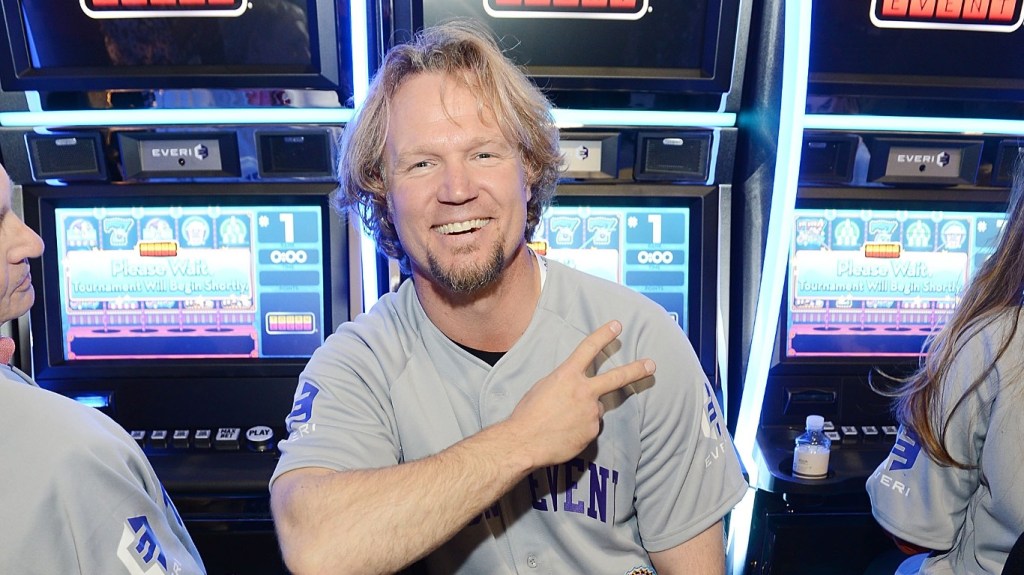 Kody Brown sitting in front of a slot machine, throwing up the peace sign and grinning