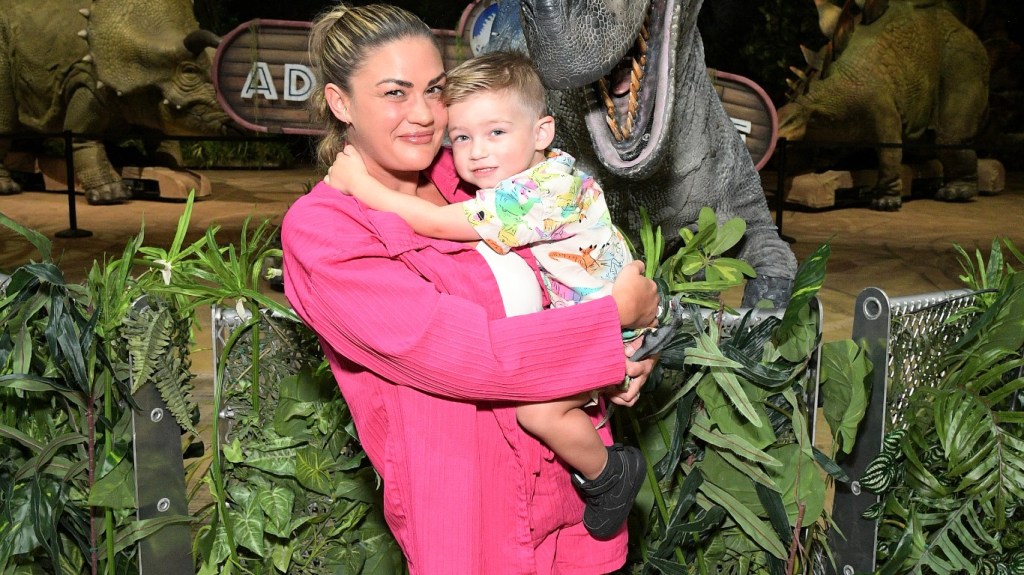 Brittany Cartwright in a pink shirt holding Cruz and standing in front of an animatronic dinosaur