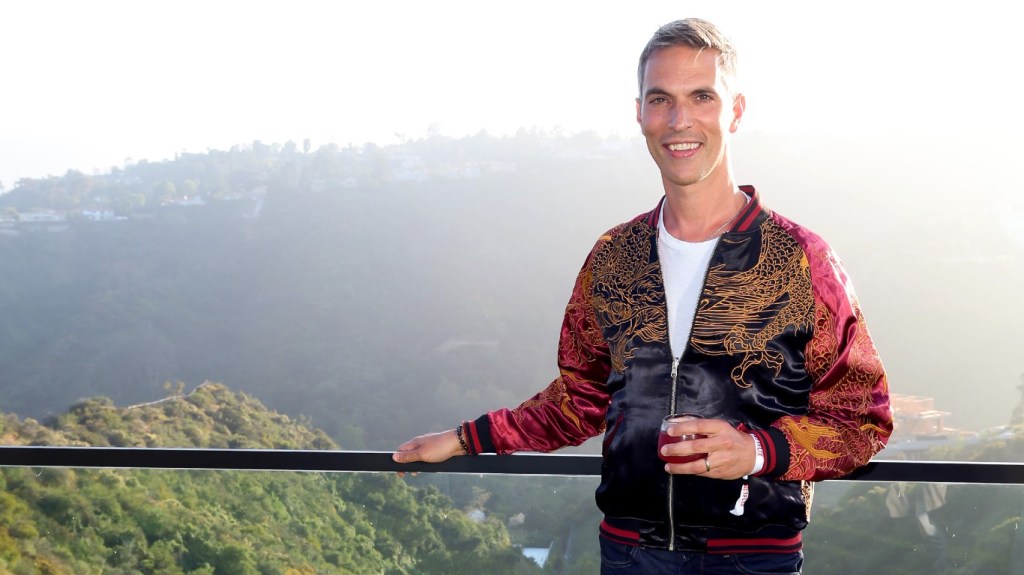 Ari Shapiro in a red and black jacket standing in front of a mountain on a balcony and holding a glass of wine