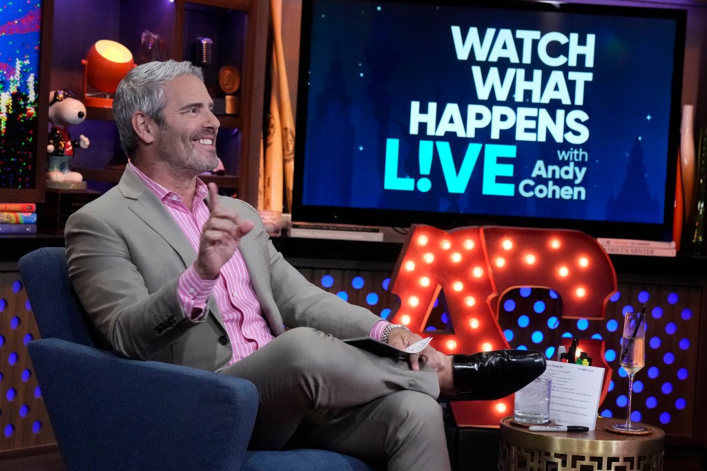 Andy Cohen in a grey suit with a pink shirt, sitting with his legs crossed on Watch What Happens Live