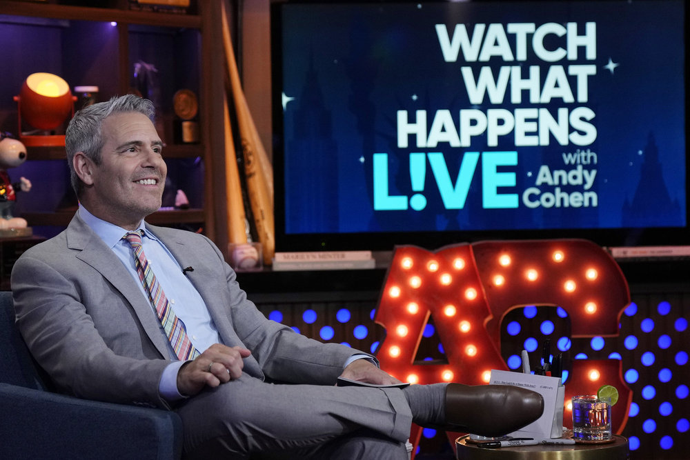 Andy Cohen in a grey suit, sitting with his legs crossed on Watch What Happens Live