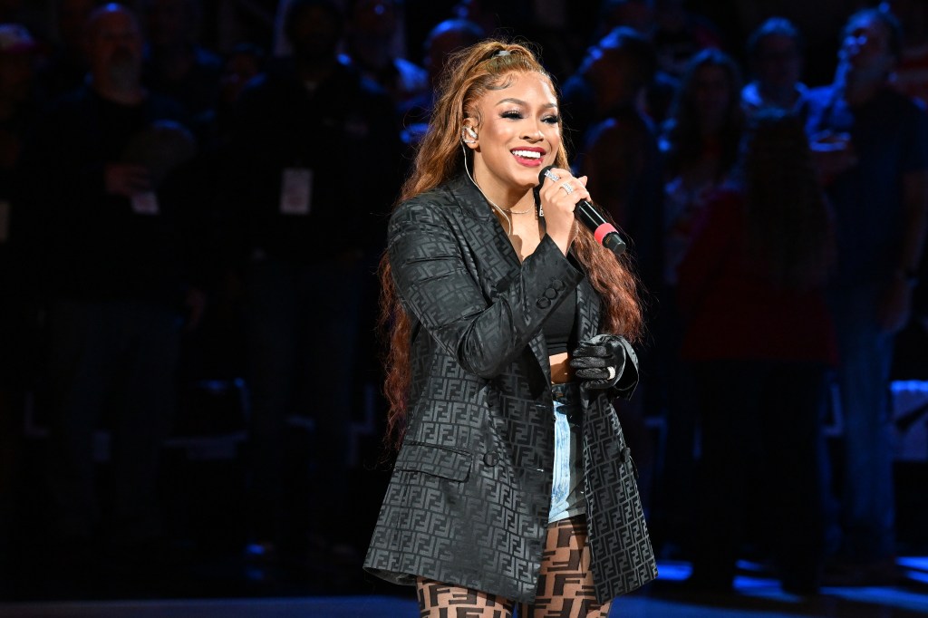 Drew Sidora dressed in a black blazer, holding a microphone, and singing during a basketball game