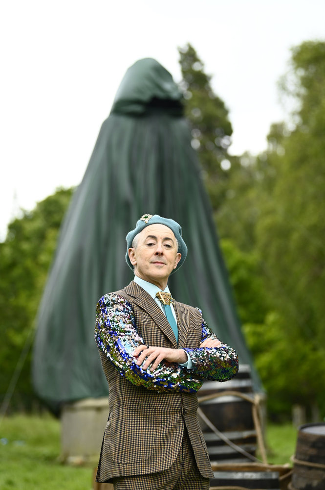 Alan Cumming posing with his arms crossed in front of a giant Traitor statue 